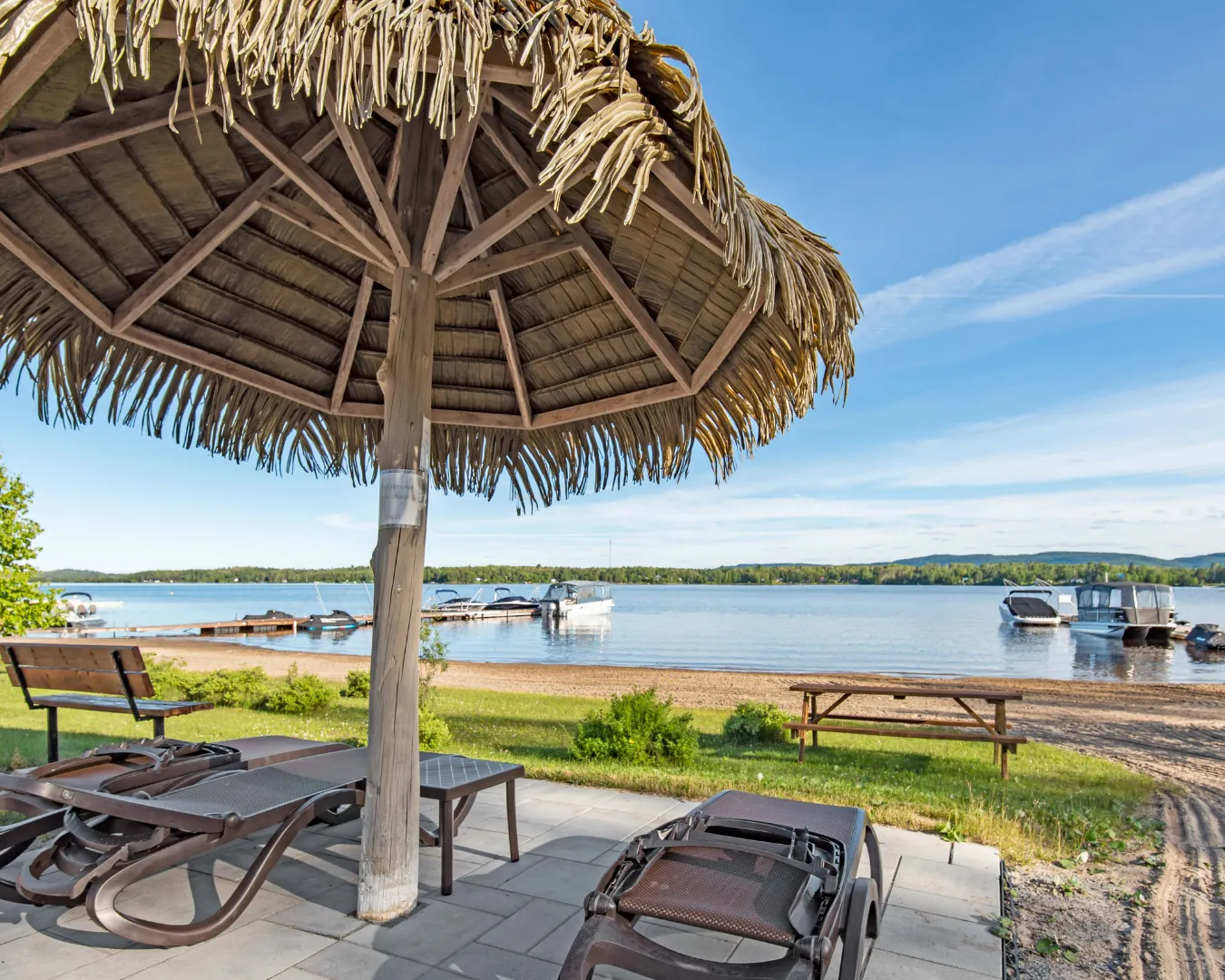 Plage au bord d’un lac avec chaises longues sous parasol en paille. Vue sur l’eau calme et quais avec bateaux.