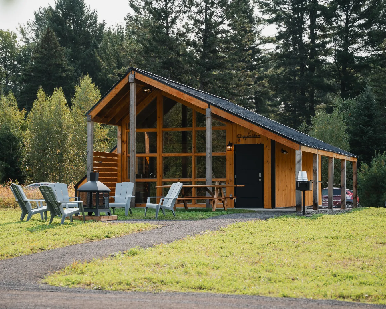 Petit chalet moderne en bois avec terrasse, foyer extérieur et chaises. Situé en pleine nature pour séjour accessible.