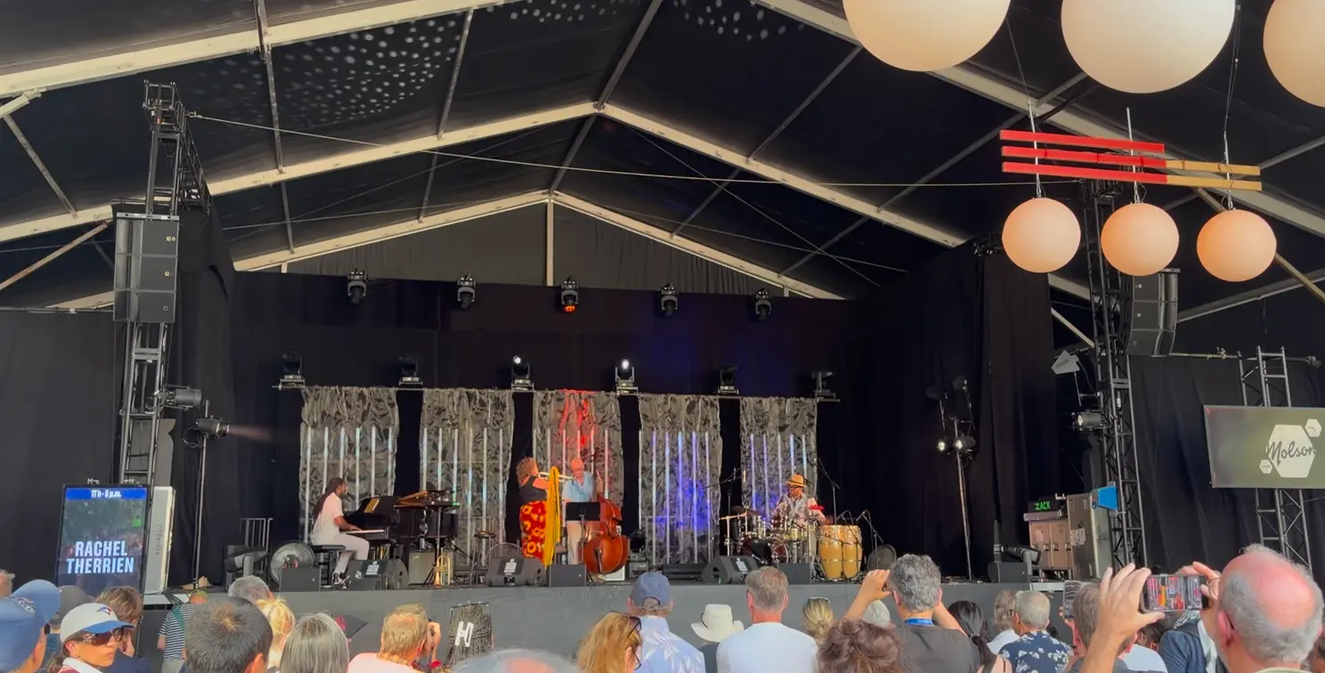 Scène de spectacle sous un chapiteau avec des musiciens en prestation et un écran latéral affichant du texte destiné au public.