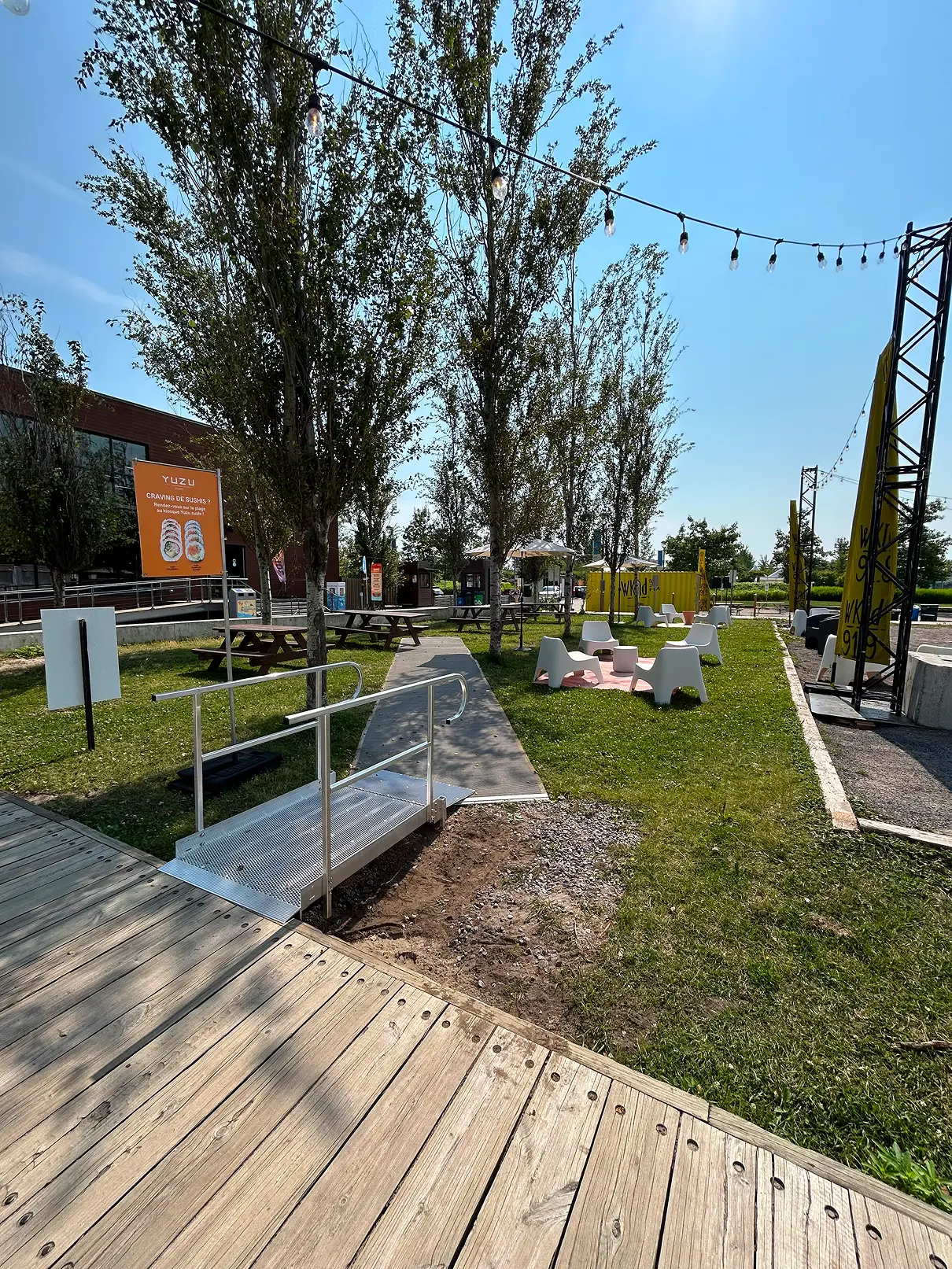 Aire de repos extérieure avec tables à pique-nique, fauteuils, cheminement aménagé et petite rampe reliant une passerelle de bois au site.