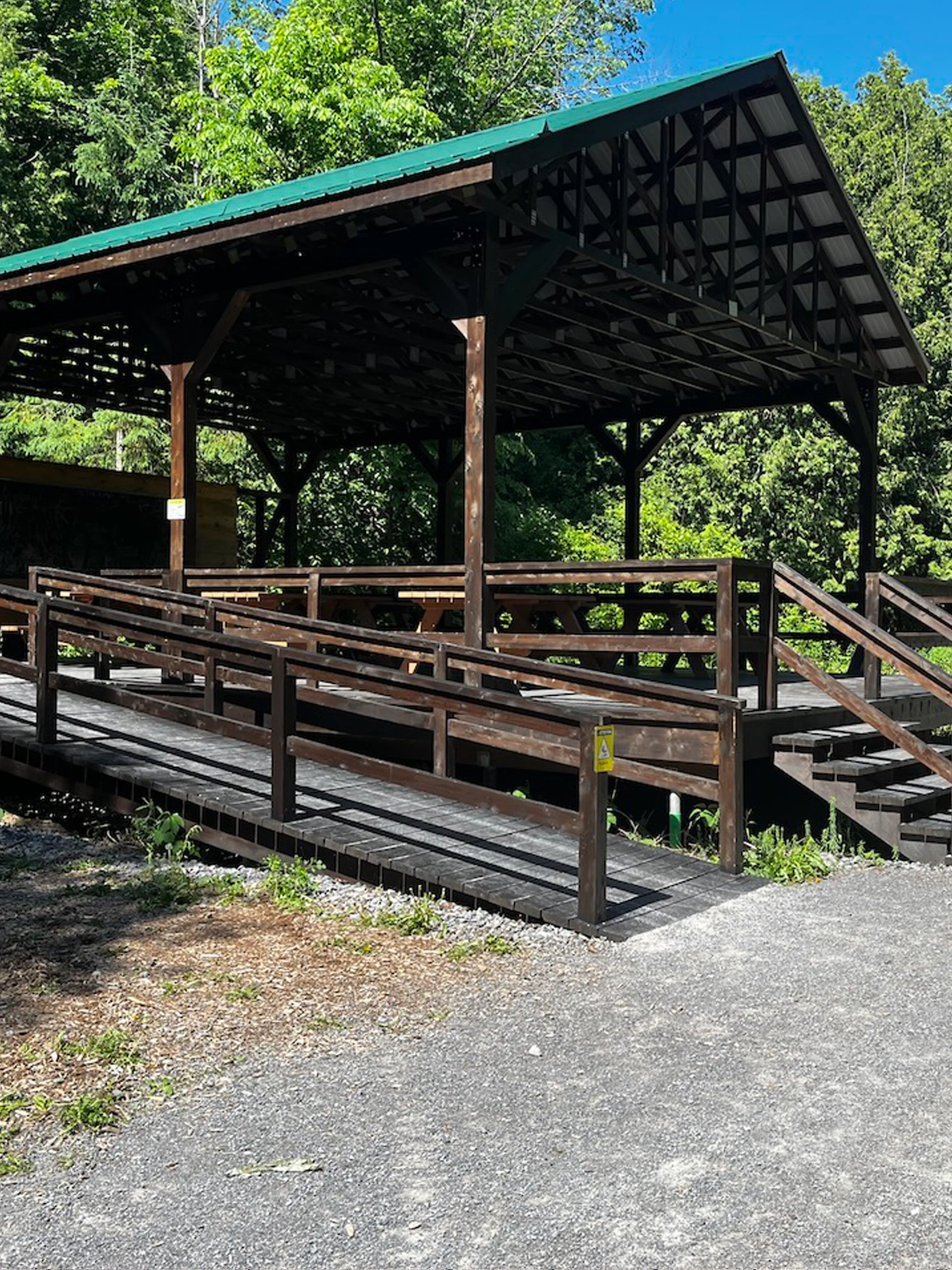 Pavillon couvert en forêt avec bancs, accessible par une rampe en bois munie de garde-corps et par un escalier.