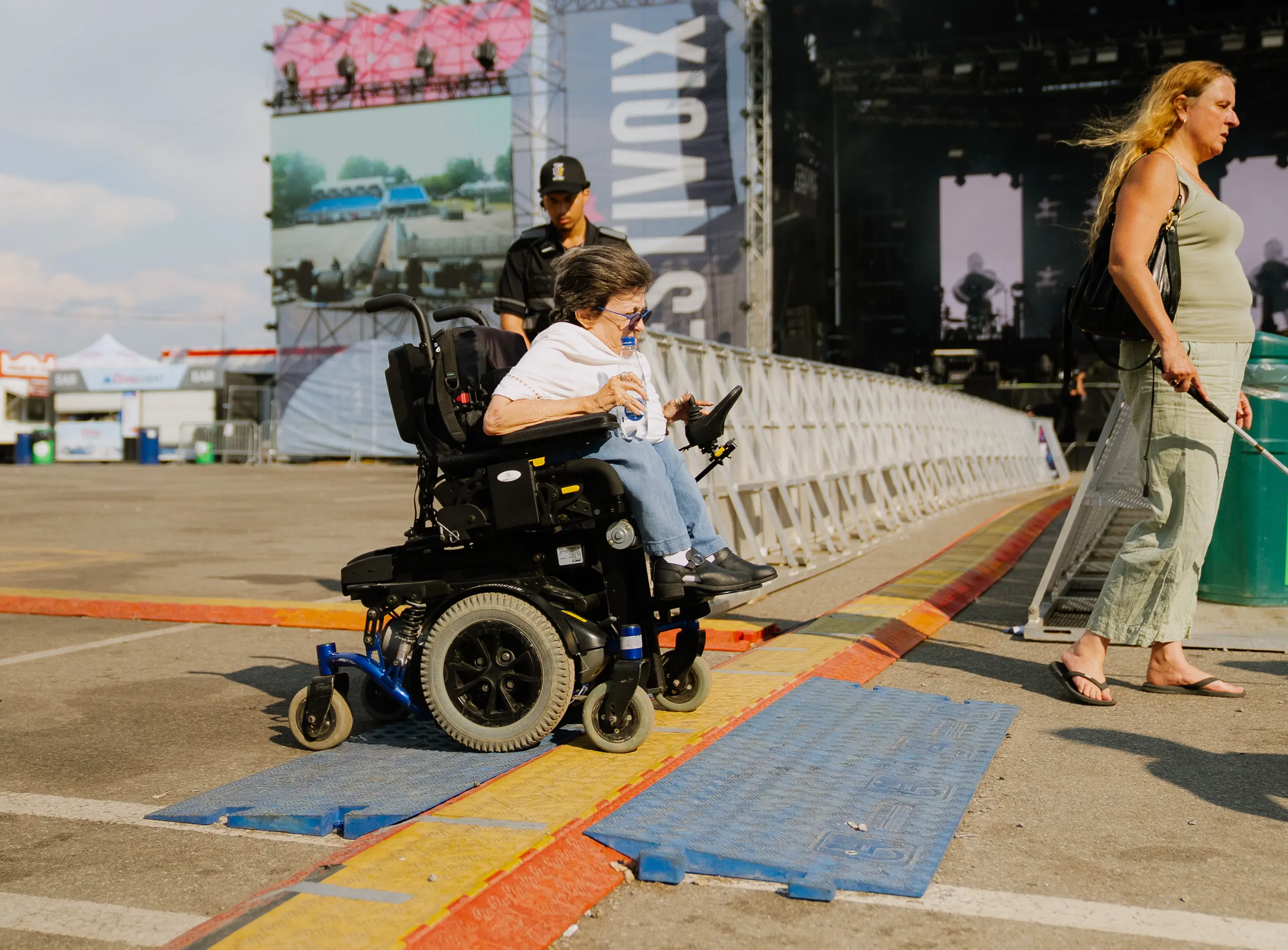 Personne en fauteuil roulant motorisé franchissant des cache-fils installés au sol devant une scène de festival.
