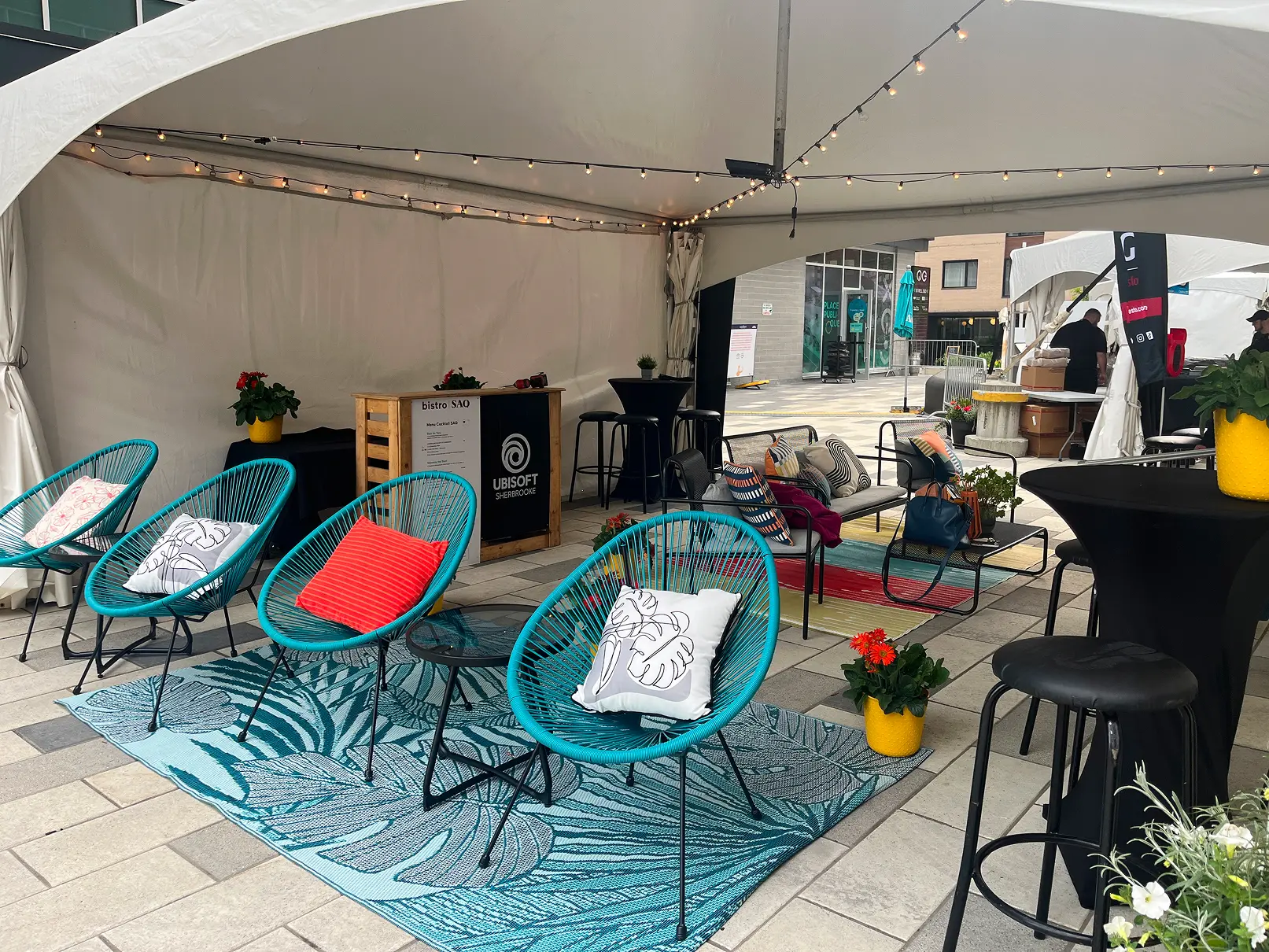 Espace couvert avec fauteuils, banquette, tables et plantes, aménagé comme zone de repos dans un festival.