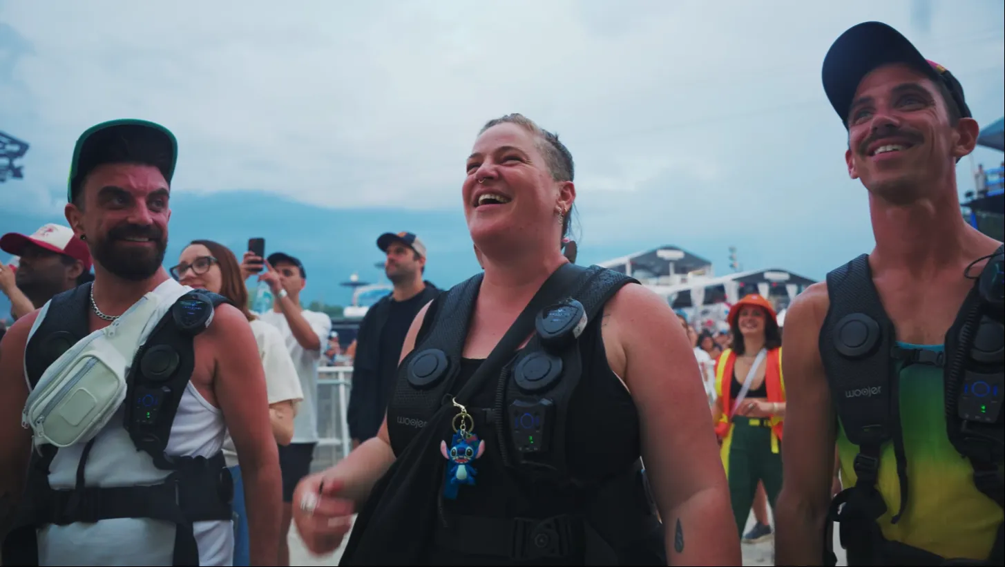 Trois festivaliers sourient devant une scène extérieure. Ils portent des gilets vibrants équipés de dispositifs tactiles sur le torse.